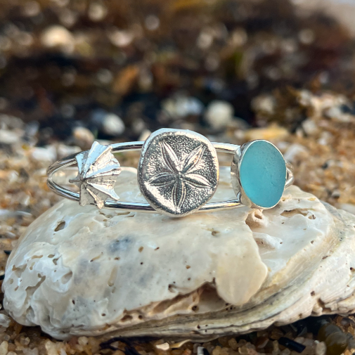 Sterling silver split centre cuff featuring a cast silver limpet shell and sea biscuit as well a blue sea glass gem. 60cm diameter with a 3cm opening.By Mornington Sea Glass
