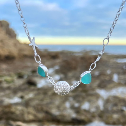 Sterling silver necklace of cast silver sea urchin shells and sea stars and blue sea glass. Hangs on a 42cm sterling silver belcher chain. By Mornington Sea Glass.